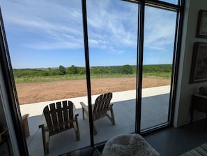 A room with large windows overlooking a dirt field and greenery, two wooden chairs, and a fluffy rug.