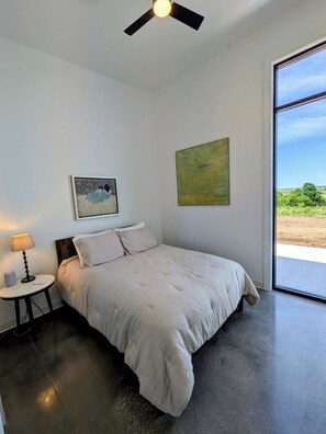 A bedroom with a bed, a ceiling fan, a nightstand with a lamp, a painting, and a large window with a view.