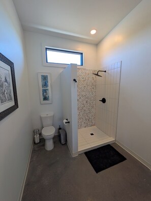 A bathroom with a toilet, a shower with a tiled wall, a small window, and a framed picture on the wall.
