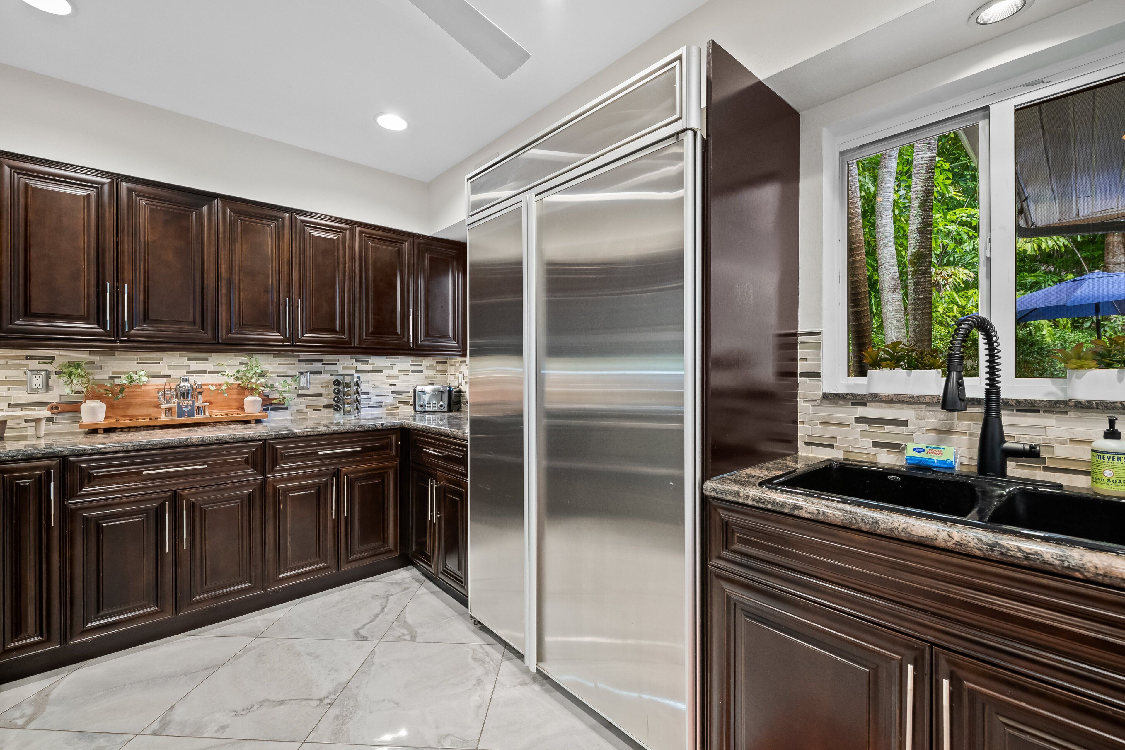 A modern kitchen with dark wood cabinets, a stainless steel refrigerator, a granite countertop, and a window with a view of trees.