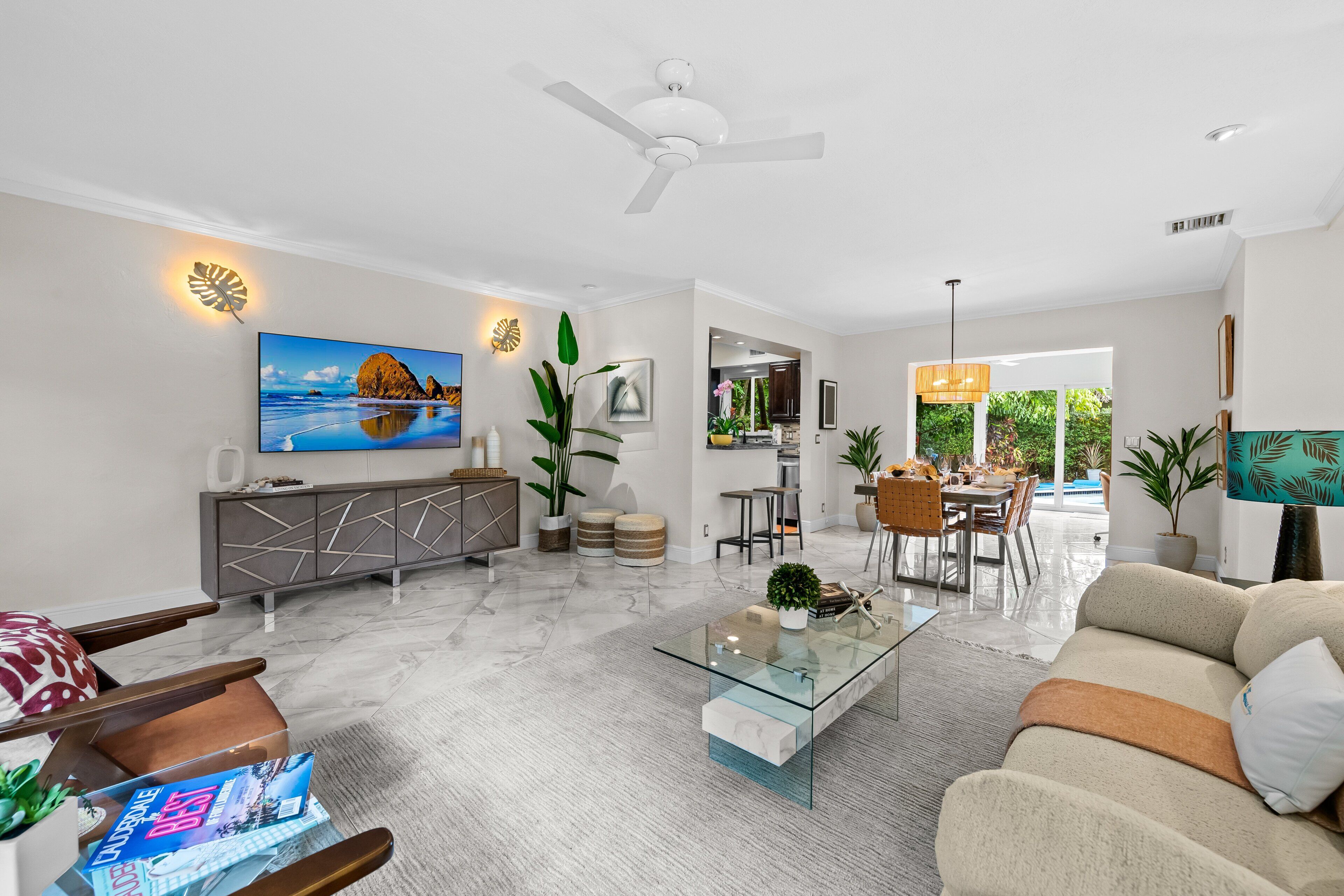 Un salon moderne avec une table basse en verre, un canapé, un coin repas avec des chaises, un ventilateur de plafond et un grand tableau accroché au mur.