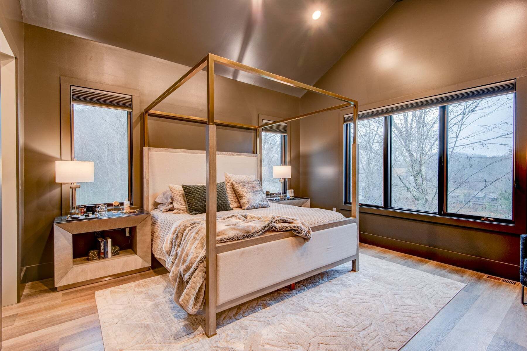 A four-poster bed with a headboard, bedside tables, and a view of trees through large windows.