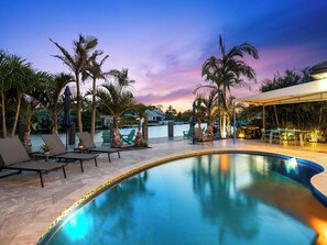 A pool area with lounge chairs and umbrellas, surrounded by palm trees and a view of a body of water at dusk. - Bahama Breeze (Oakland Park)
