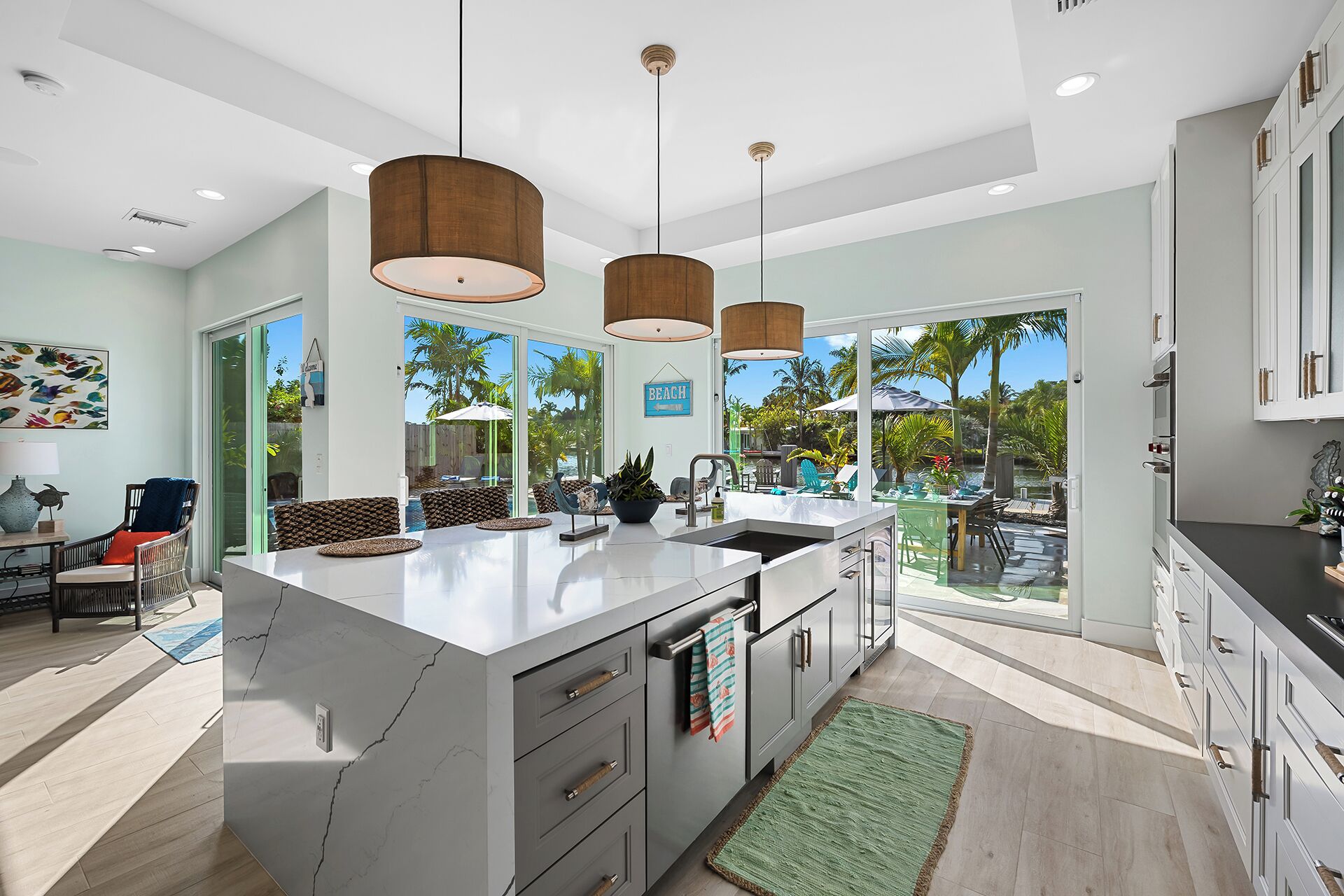 A modern kitchen with a large island, pendant lights, and a view of a pool and palm trees.