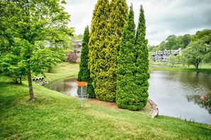 A serene park with a pond, neatly trimmed hedges, and a disc golf basket.
