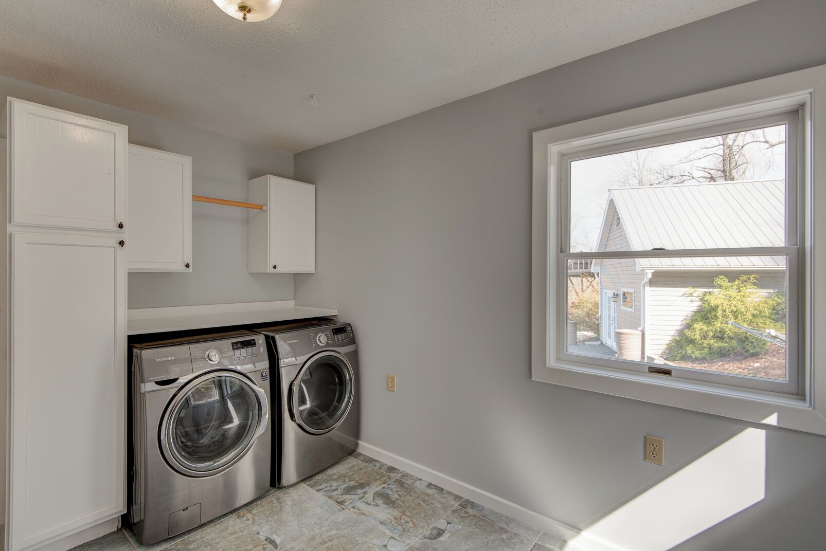 A laundry room with two front-loading washers, a window with a view of a house, and white cabinetry.