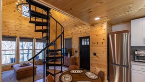 A wooden cabin interior with a spiral staircase, a dining table with coasters, a kitchen with stainless steel appliances, and a black door. - Retreat at Riverbend (West Jefferson)