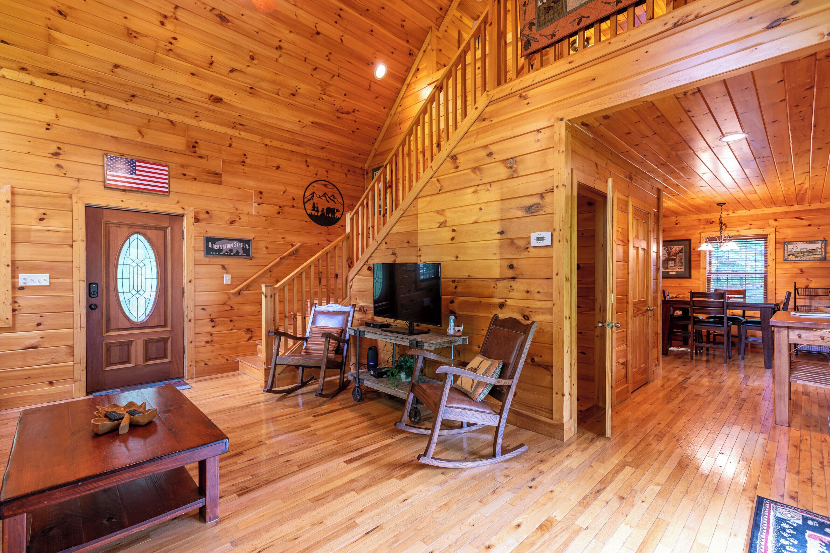 A wooden cabin interior with a dining area, a television, rocking chairs, and a staircase.