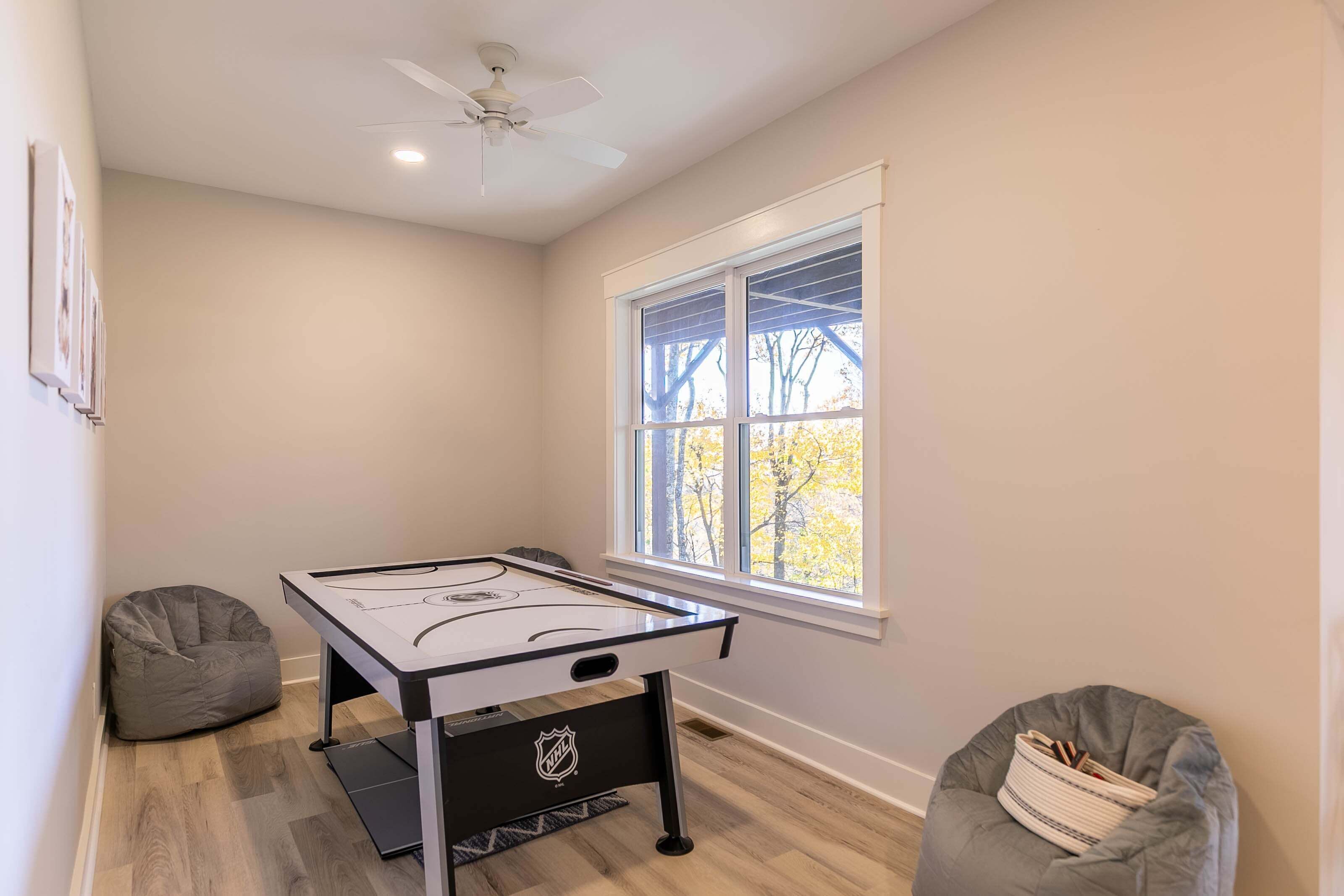 A room with a pool table, two bean bags, a ceiling fan, and a window with a view of trees.