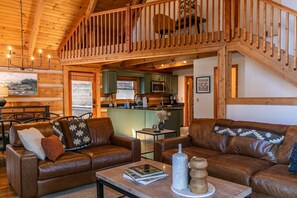 A cozy cabin interior with a living room, dining area, and kitchen, featuring wooden beams and a loft bedroom.