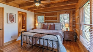 Quarto de cabana de madeira com cama, duas mesas de cabeceira, ventoinha no teto e janela com persianas.
