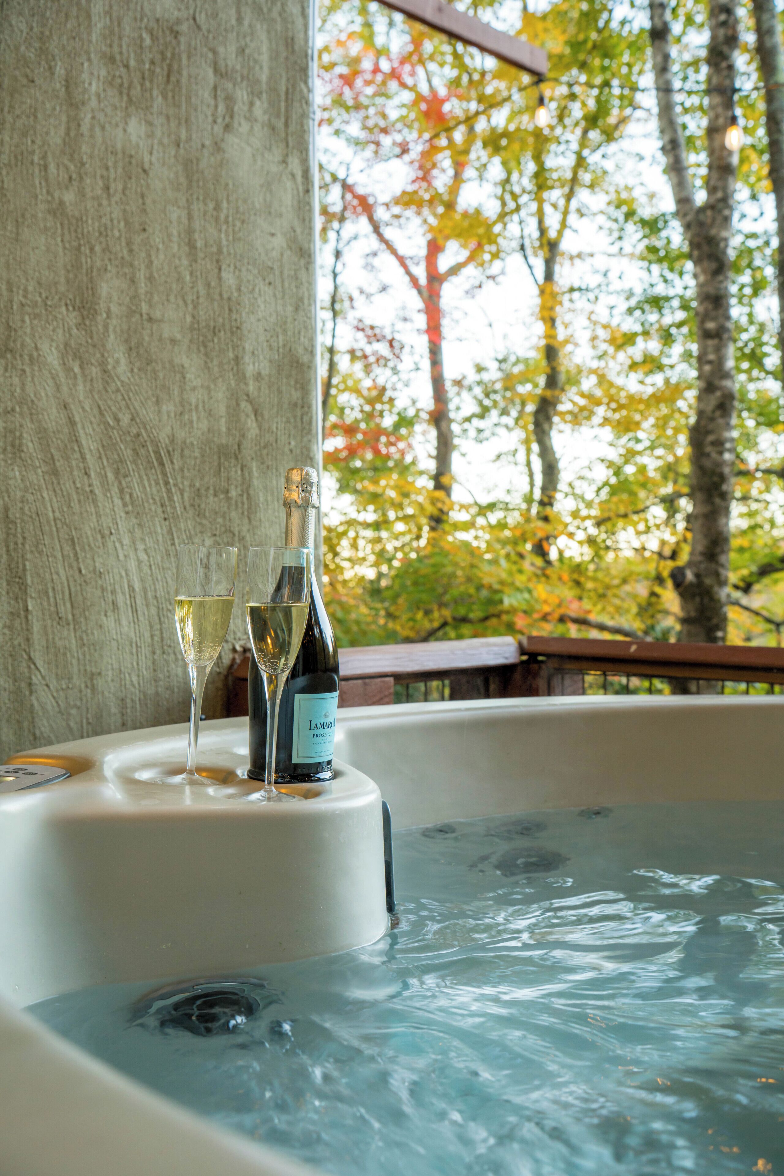 Un bain à remous avec une bouteille de champagne et un verre, entouré d’une terrasse en bois et d’arbres.