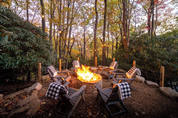 A circular outdoor seating area with a fire pit surrounded by wooden chairs, set in a forested environment.
