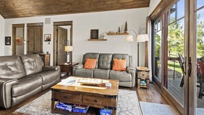 A living room with a leather sofa, a wooden coffee table, and a grey couch with orange cushions. There are wooden shelves with decorative items and a large window providing a view of the outdoors. - Sommer Hus at Eagles Nest (Elk Park)