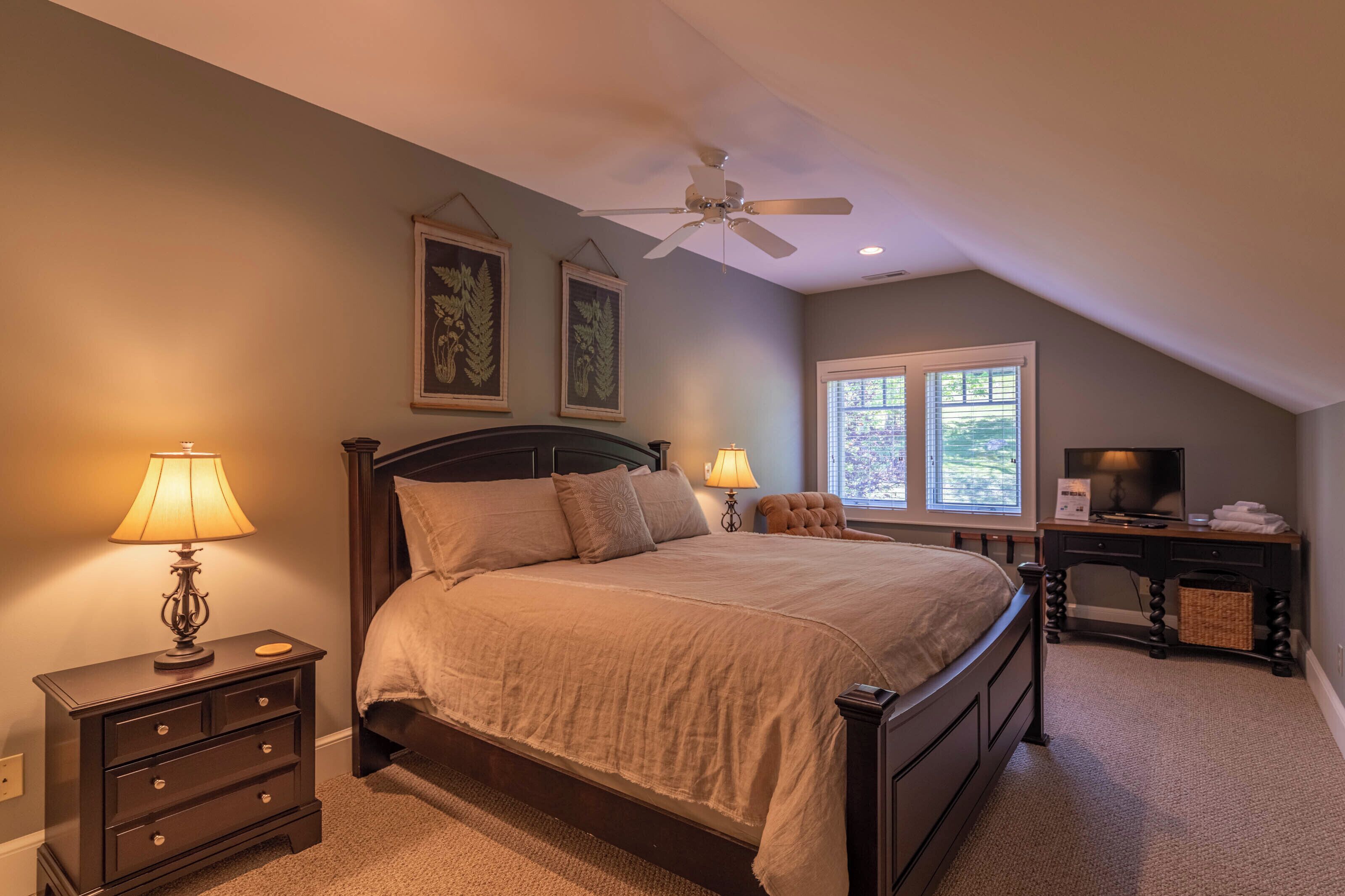 A bedroom with a bed, nightstands, a desk, a ceiling fan, and windows.
