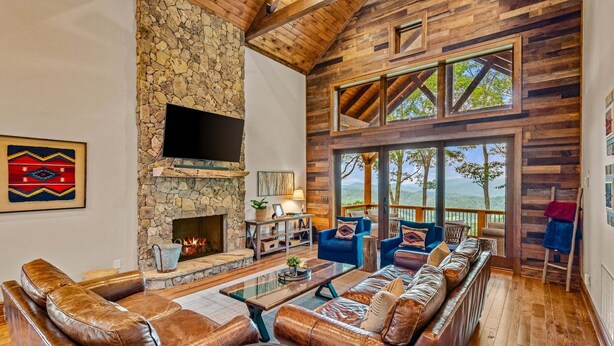 A spacious living room with a stone fireplace, leather sofas, and large windows offering a view of the outdoors. - Blue Ridge View at Eagles Nest (Banner Elk)