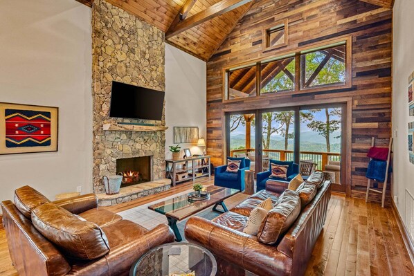 Ein geräumiges Wohnzimmer mit Kamin aus Stein, Ledersofas und großen Fenstern mit Blick ins Freie.