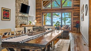 A rustic interior with a stone fireplace, wooden dining table, and large windows offering a view of the outdoors. - Blue Ridge View at Eagles Nest (Banner Elk)
