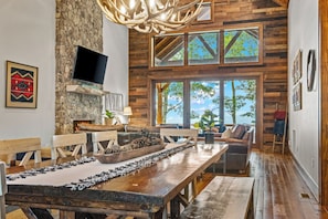 Ein rustikales Interieur mit Kamin aus Stein, Esstisch aus Holz und großen Fenstern mit Blick ins Freie.
