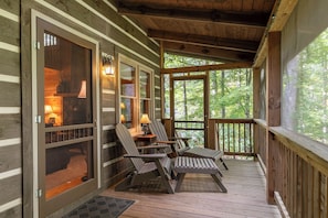 A wooden deck with Adirondack chairs and a table, a door leading to a room with a lamp, and a view of trees through the door. - 1 Sweet Seclusion Peaceful Cabin Just Outside of Boone With hot tub (Vilas)