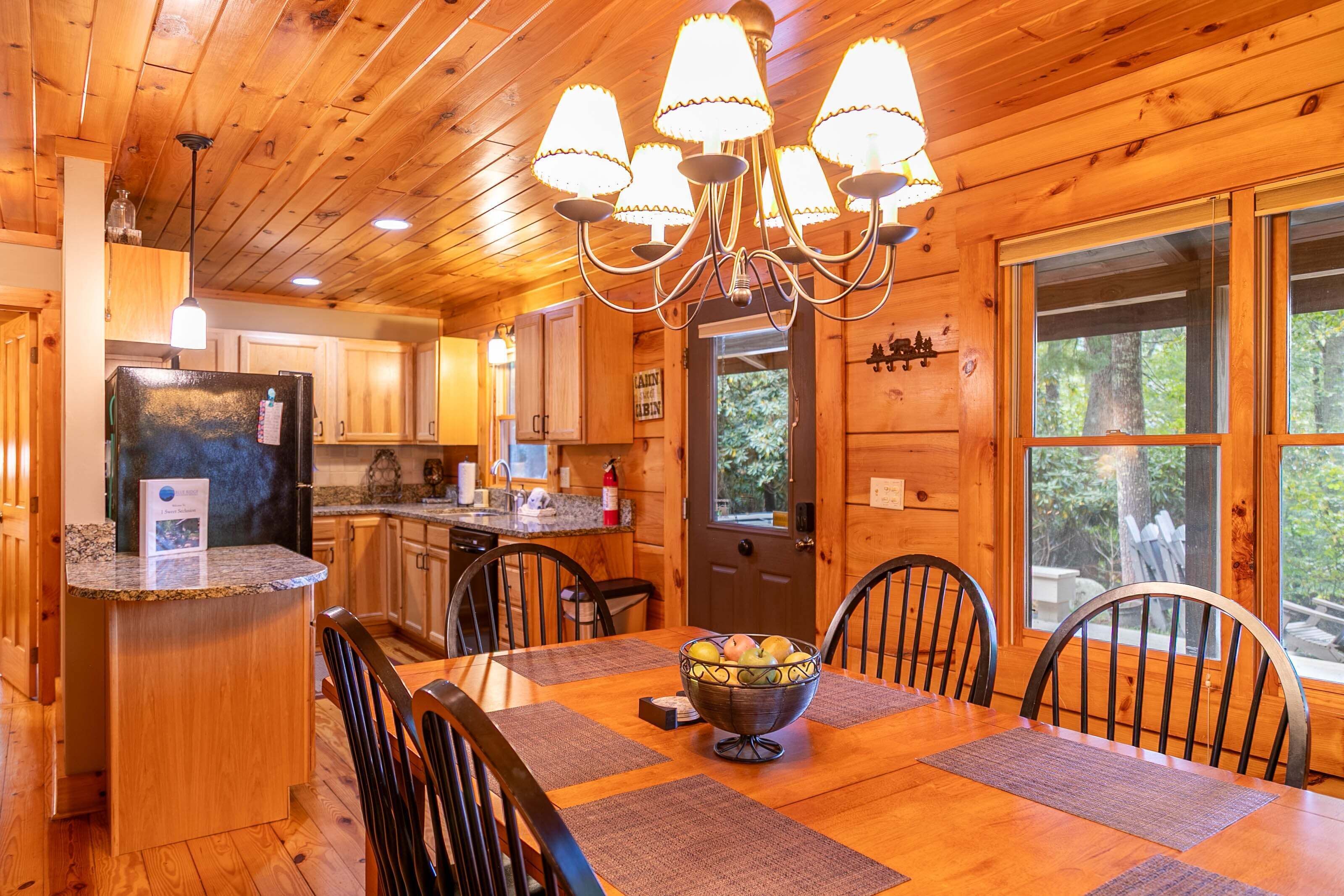 A wooden cabin interior with a dining table, chairs, a chandelier, a refrigerator, and a window with a view of trees.