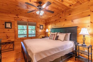 A wooden cabin bedroom with a bed, two bedside lamps, a ceiling fan, and a window with a view of trees. - 1 Sweet Seclusion Peaceful Cabin Just Outside of Boone With hot tub (Vilas)