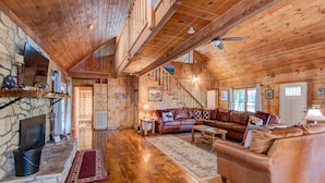 A log cabin interior with a stone fireplace, wooden floors, a leather sofa, and a staircase. - Hidden Serenity Hot Tub, Fire Pit, Minutes From Boone (Fleetwood)