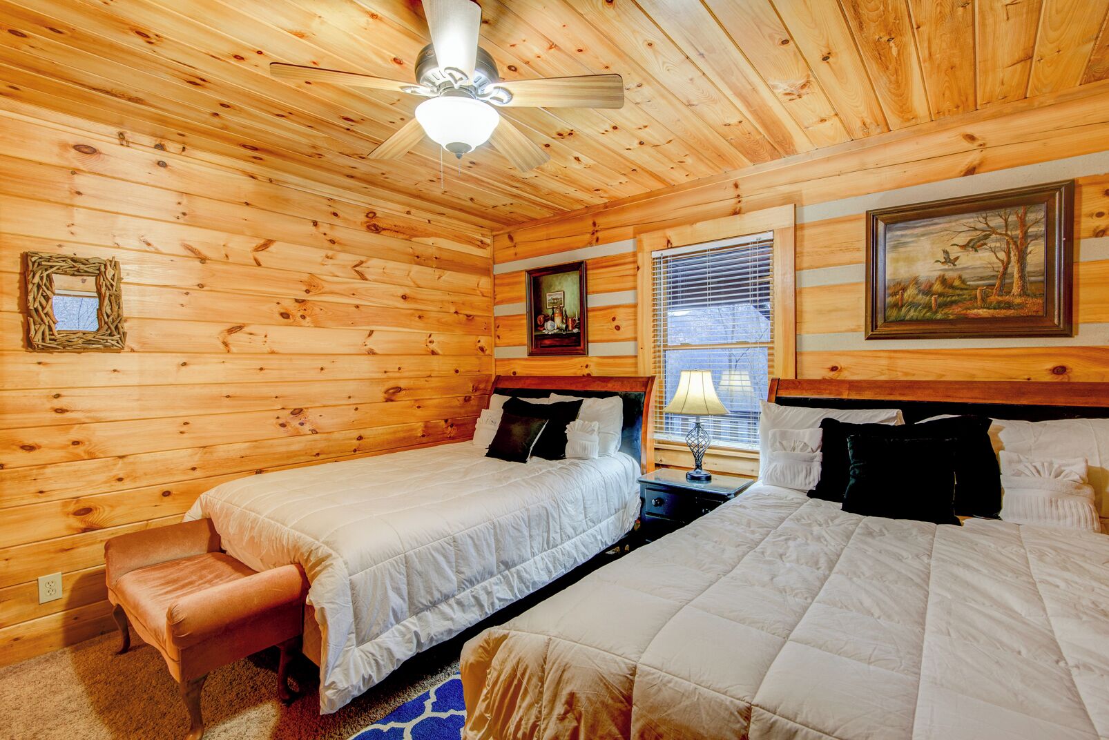 A log cabin room with two beds, a ceiling fan, a window with blinds, and a painting on the wall.