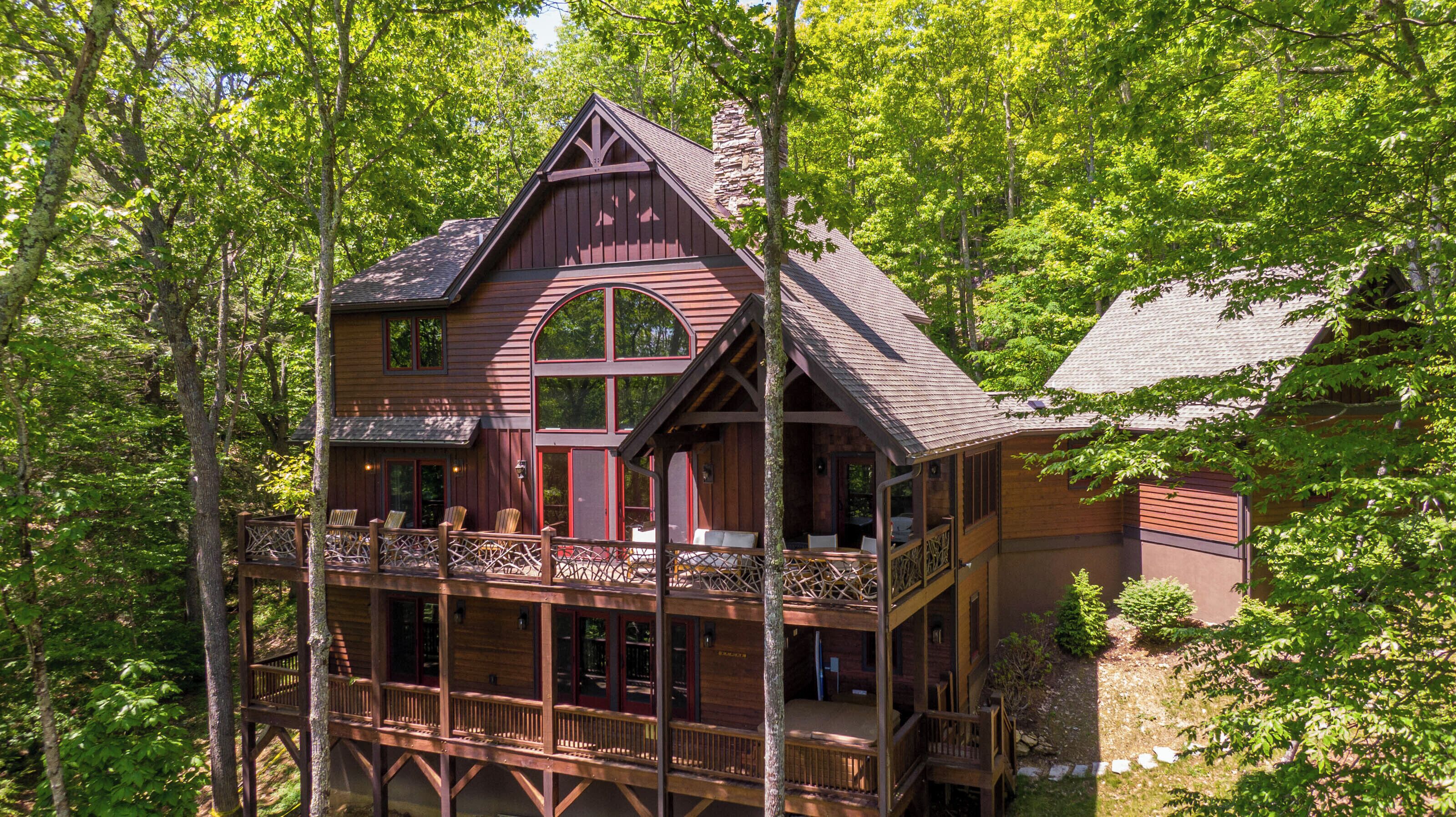A two-story log cabin with a balcony and large windows, surrounded by dense forest.
