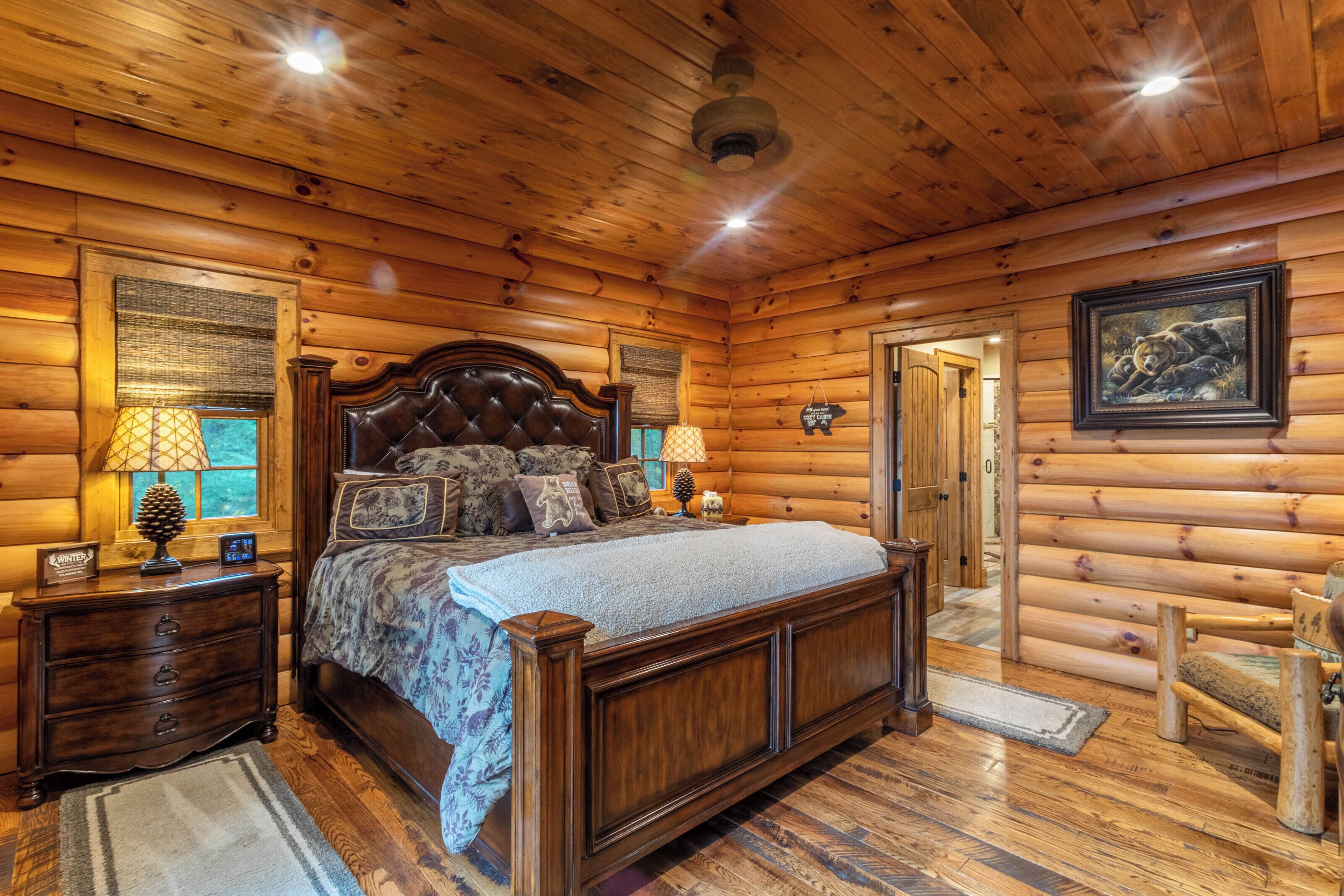 Un dormitorio de cabaña de troncos con una cama grande, cabecera de madera, dos mesitas de noche con lámparas, una silla de madera y un cuadro en la pared.