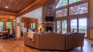 A log cabin interior with a stone fireplace, large windows, and a cozy seating area. - Eagles Landing at Eagles Nest (Banner Elk)
