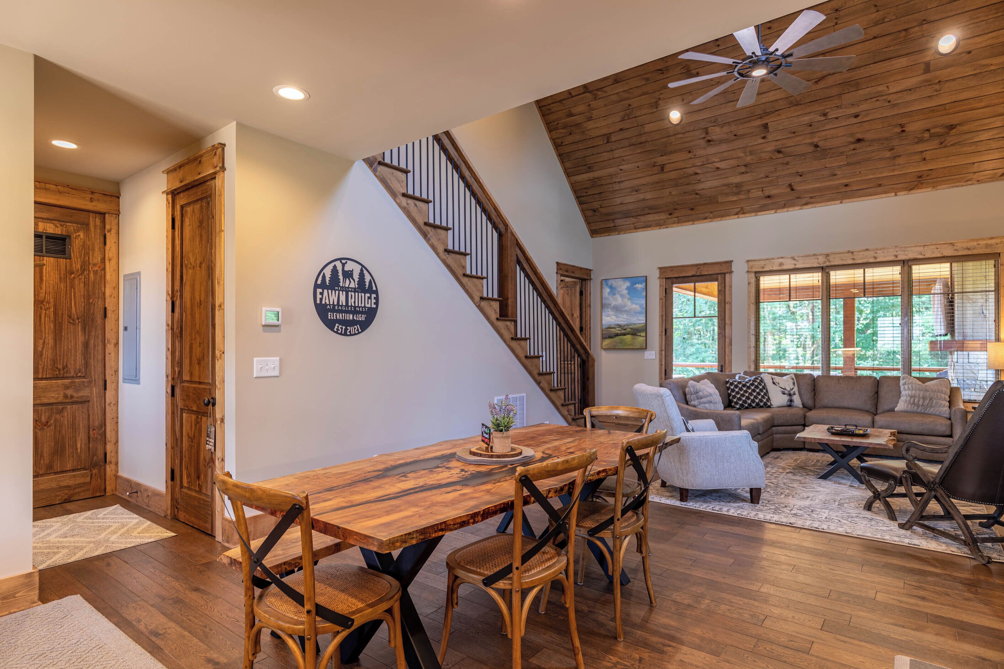 A spacious living area with a wooden staircase, a dining table with chairs, and a large window providing a view of the outdoors.
