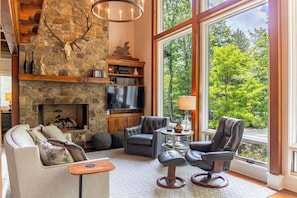 Ein gemütliches Wohnzimmer mit Kamin aus Stein, Ledermöbeln und einem großen Fenster mit Blick auf den Wald.