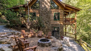 A rustic cabin with a stone chimney, wooden deck, Adirondack chairs, and a fire pit.