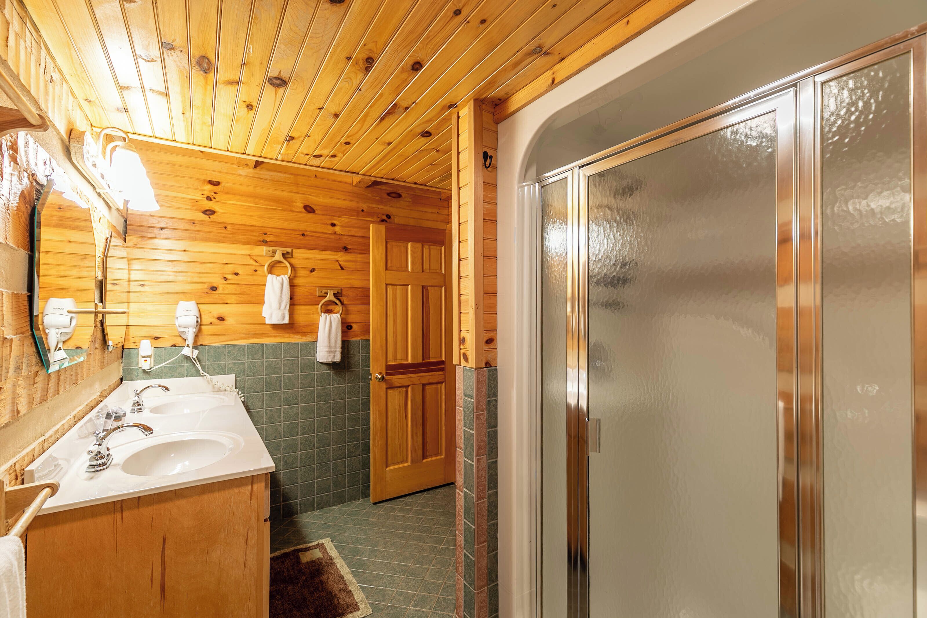 A wooden cabin bathroom with a wooden vanity, two sinks, a mirror, and a shower stall.
