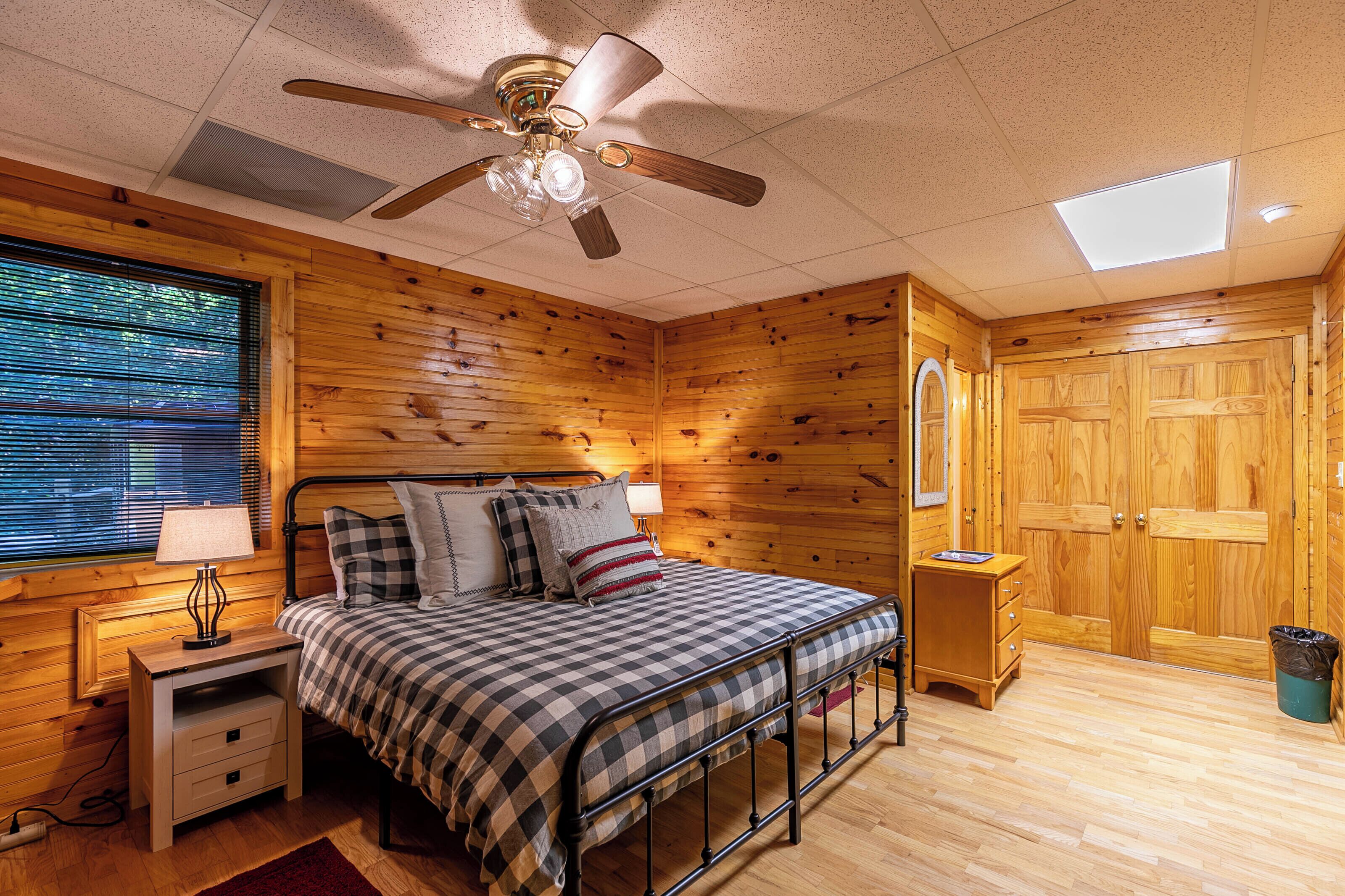 A bedroom with a bed, a nightstand, a ceiling fan, a window with blinds, and wooden walls.