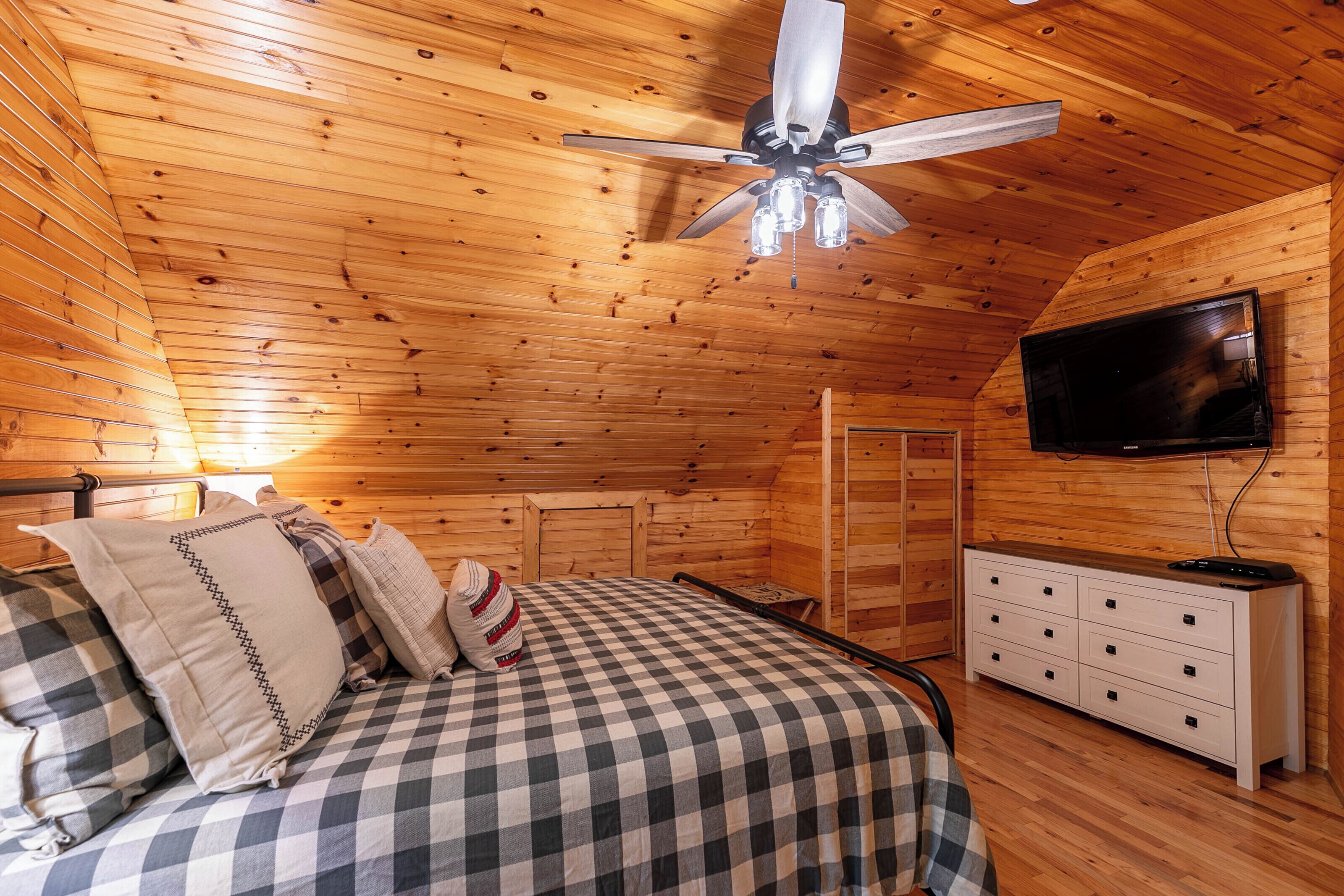 Un dormitori d'una cabana de fusta amb un llit, una petita taula de nit, televisió i ventilador de sostre.