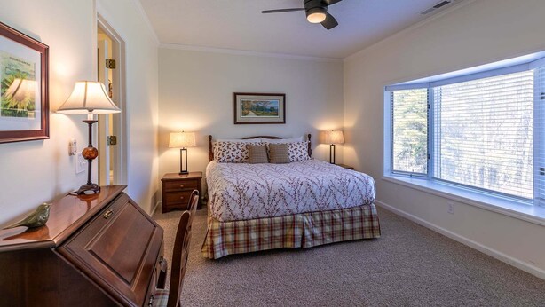 A bedroom with a bed, two bedside lamps, a ceiling fan, a window with blinds, and a framed picture on the wall.