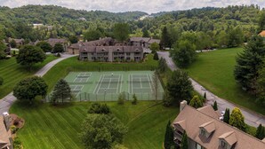 A large building with a tennis court, surrounded by lush greenery and a hilly landscape.