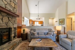A cozy living room with a stone fireplace, a grey sofa, a wooden coffee table, and a kitchen in the background.