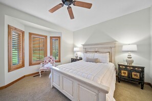A bedroom with a large bed, a ceiling fan, wooden shutters, a nightstand, and a chair. - Chetola Sierra 1 (Blowing Rock)