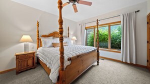 A four-poster bed with a ceiling fan, a nightstand with a lamp, a window with curtains, and a view of greenery outside.
