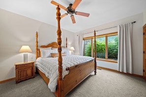 Un lit à baldaquin avec un ventilateur de plafond, une table de chevet avec une lampe, une fenêtre avec des rideaux et une vue sur la verdure à l’extérieur.