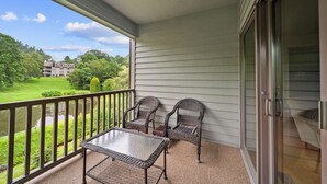 A balcony with a table and two chairs, a glass door, and a view of a building and greenery.