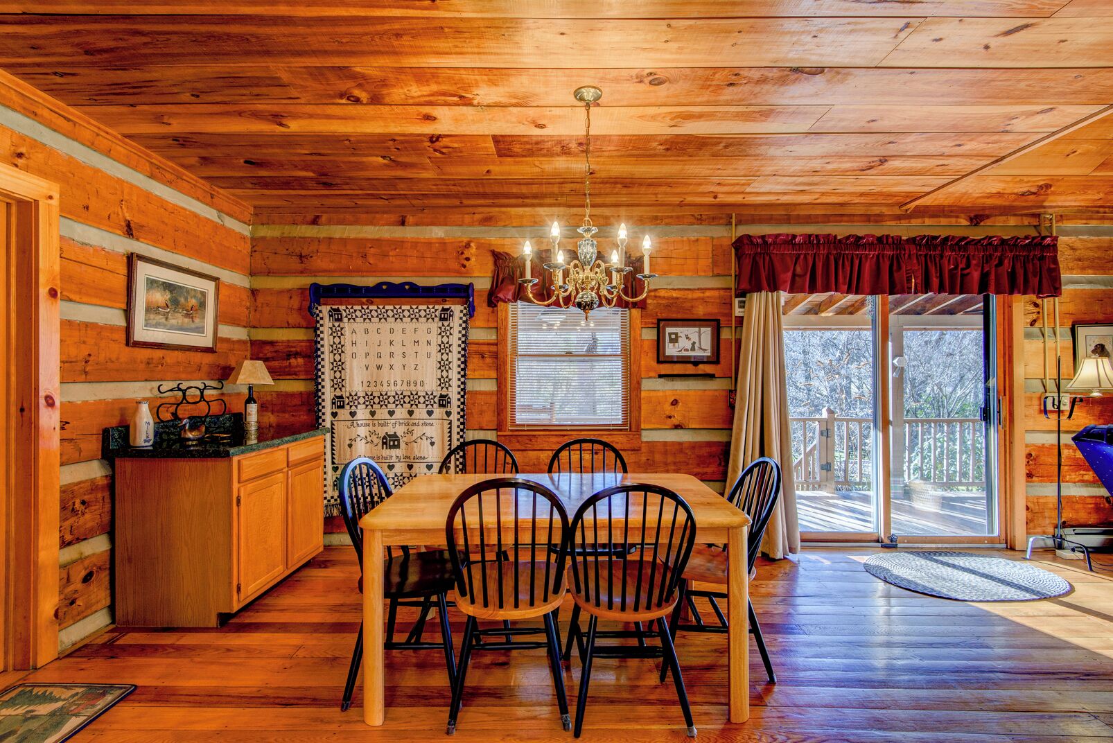 A wooden cabin interior with a dining table, chairs, a chandelier, and a kitchen area.