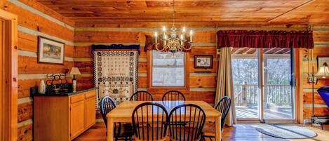 A wooden cabin interior with a dining table, chairs, a chandelier, and a kitchen area.