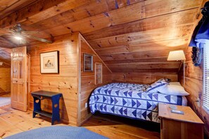 Un dormitorio de cabaña de madera con una cama, una mesita pequeña, un ventilador de techo y cuadros en las paredes.