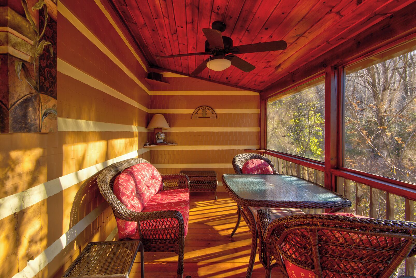 A cozy cabin interior with a wooden ceiling, a ceiling fan, wicker furniture, and a large window offering a view of trees.