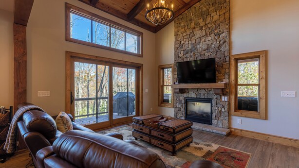 A spacious living room with a stone fireplace, leather sofas, and large windows offering a view of the outdoors. - Hidden Gem at Eagles Nest (Banner Elk)