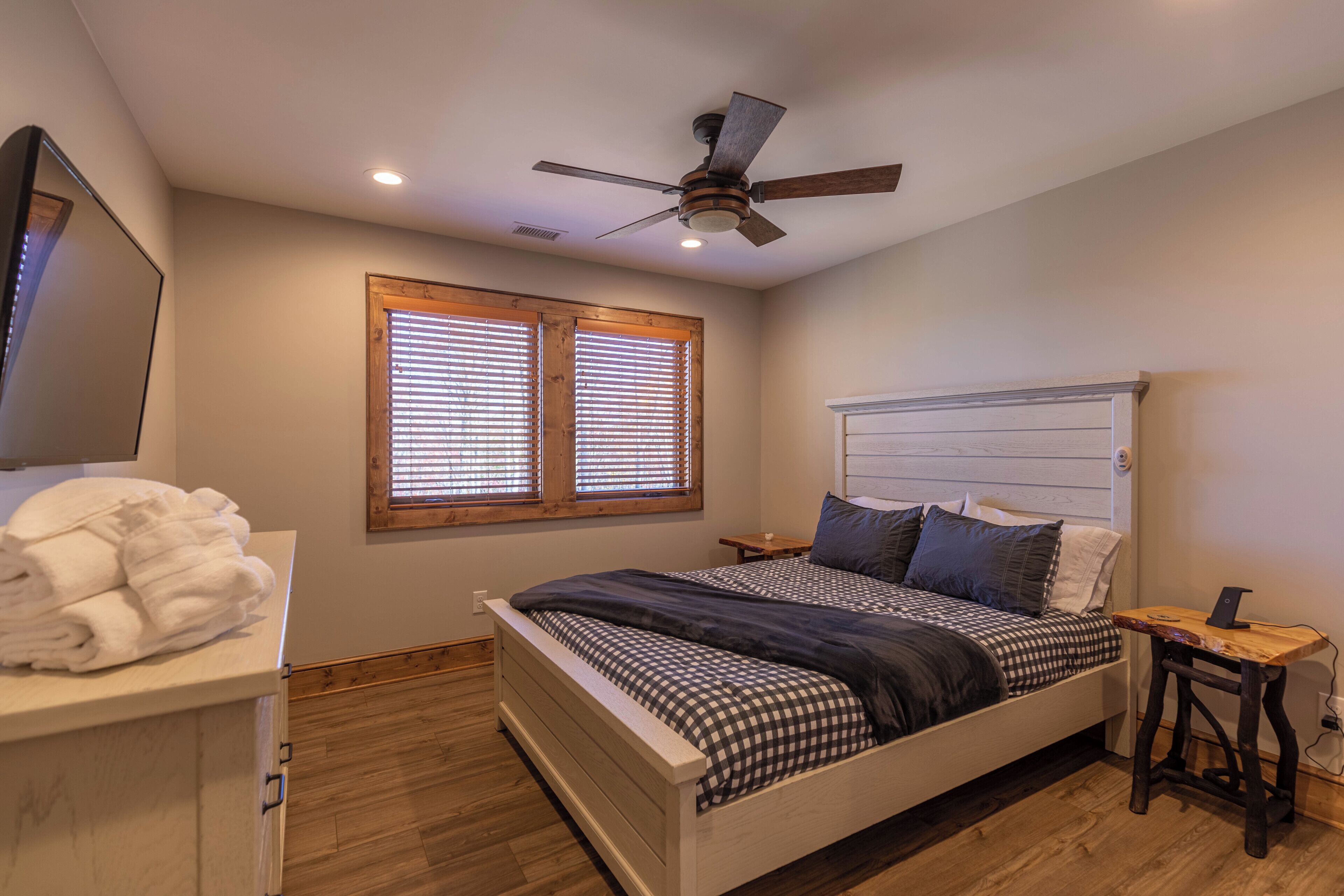 A bedroom with a bed, a ceiling fan, wooden blinds, a bedside table, and a lamp.